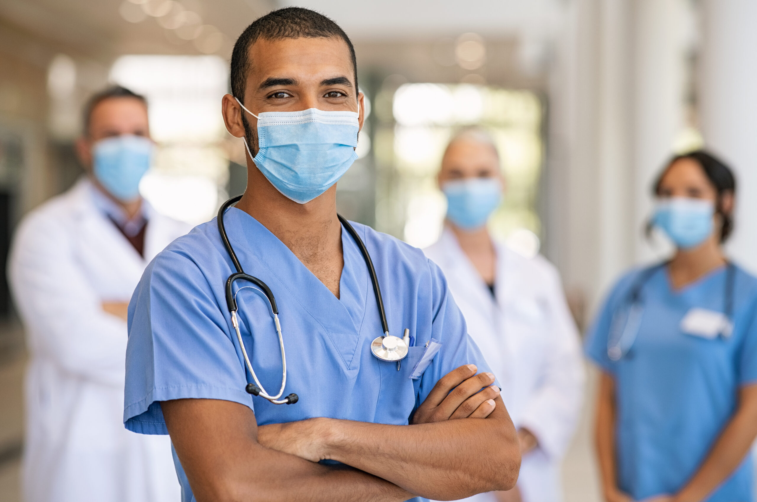 Confident multiethnic male nurse in front of his medical team looking at camera wearing face mask during covid-19 outbreak. Happy and proud indian young surgeon standing in front of his colleagues.